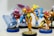 A collection of colorful figurines, with a focus on a central character in orange and yellow armor holding a blue weapon. Surrounding figures are similarly stylized with vibrant colors and dynamic poses, all standing on display bases.