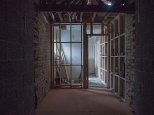 A dimly lit construction area with exposed brick walls and wooden framing. The space appears to be undergoing renovations, with tools and equipment visible in the background. Light enters from the left, highlighting a partially constructed wall.