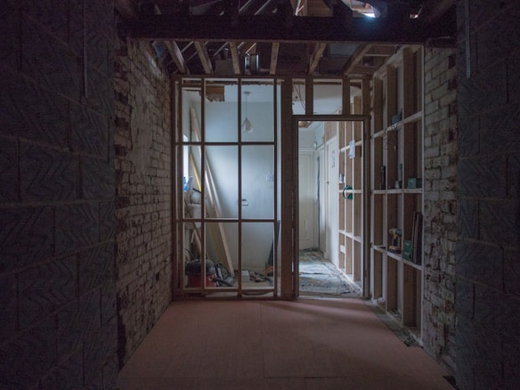 A dimly lit construction area with exposed brick walls and wooden framing. The space appears to be undergoing renovations, with tools and equipment visible in the background. Light enters from the left, highlighting a partially constructed wall.