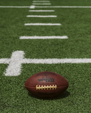 An American football rests on a grassy field, centered near white markings that indicate yard lines.