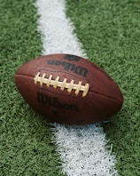 A close-up of an American football placed on a green artificial turf field. The football is positioned across a white line on the field, with the logo displayed prominently.