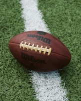 A close-up of an American football placed on a green artificial turf field. The football is positioned across a white line on the field, with the logo displayed prominently.