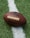 A close-up of an American football placed on a green artificial turf field. The football is positioned across a white line on the field, with the logo displayed prominently.