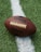 A close-up of an American football placed on a green artificial turf field. The football is positioned across a white line on the field, with the logo displayed prominently.