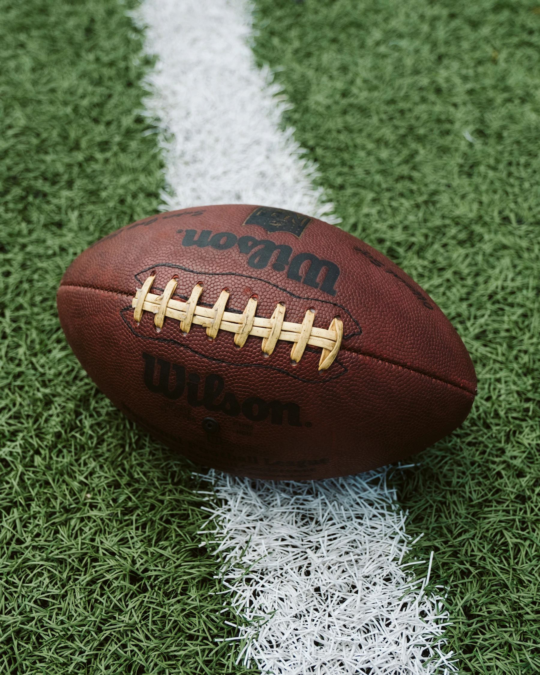 Close-up of a brown leather football's white laces and pebbled cover, lying across a white line on turf