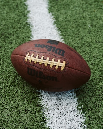 A close-up of an American football placed on a green artificial turf field. The football is positioned across a white line on the field, with the logo displayed prominently.