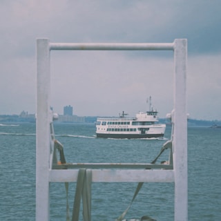 A ferry labeled 'Statue Cruises' is sailing on the water with a cloudy sky and city buildings in the distance. The ferry is framed by metal structures in the foreground.