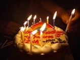 A close-up of a birthday cake with a rainbow decoration and lit candles. The cake features colorful icing with a message in red lettering. Hands are visible lighting the candles.