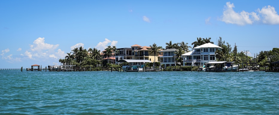 A row of beachfront houses lined along a tranquil body of water, with palm trees scattered among the properties. The sky is mostly clear with a few scattered clouds, and the water is calm with small waves lapping gently against the shore.