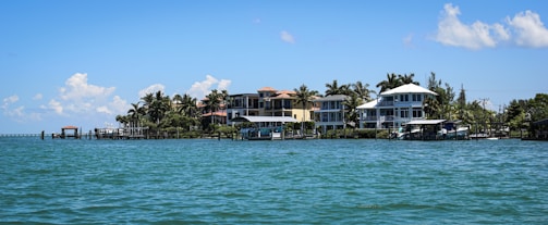 A row of beachfront houses lined along a tranquil body of water, with palm trees scattered among the properties. The sky is mostly clear with a few scattered clouds, and the water is calm with small waves lapping gently against the shore.