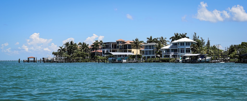 A row of beachfront houses lined along a tranquil body of water, with palm trees scattered among the properties. The sky is mostly clear with a few scattered clouds, and the water is calm with small waves lapping gently against the shore.