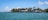 A row of beachfront houses lined along a tranquil body of water, with palm trees scattered among the properties. The sky is mostly clear with a few scattered clouds, and the water is calm with small waves lapping gently against the shore.