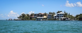A row of beachfront houses lined along a tranquil body of water, with palm trees scattered among the properties. The sky is mostly clear with a few scattered clouds, and the water is calm with small waves lapping gently against the shore.