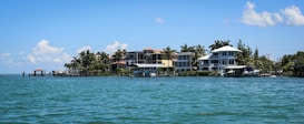 A row of beachfront houses lined along a tranquil body of water, with palm trees scattered among the properties. The sky is mostly clear with a few scattered clouds, and the water is calm with small waves lapping gently against the shore.