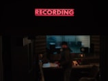 A recording studio is depicted with a prominent red 'RECORDING' sign illuminated at the top. Below, a person stands near a mixing console, surrounded by audio equipment and speakers. The environment is dimly lit, suggesting an atmosphere of focus and creativity.