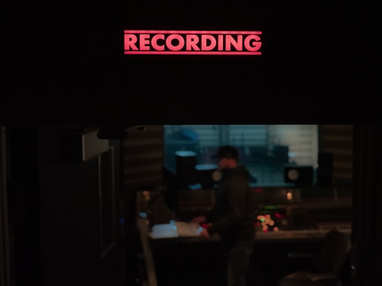 A recording studio is depicted with a prominent red 'RECORDING' sign illuminated at the top. Below, a person stands near a mixing console, surrounded by audio equipment and speakers. The environment is dimly lit, suggesting an atmosphere of focus and creativity.