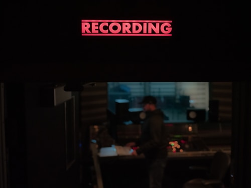 A recording studio is depicted with a prominent red 'RECORDING' sign illuminated at the top. Below, a person stands near a mixing console, surrounded by audio equipment and speakers. The environment is dimly lit, suggesting an atmosphere of focus and creativity.