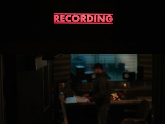 A recording studio is depicted with a prominent red 'RECORDING' sign illuminated at the top. Below, a person stands near a mixing console, surrounded by audio equipment and speakers. The environment is dimly lit, suggesting an atmosphere of focus and creativity.