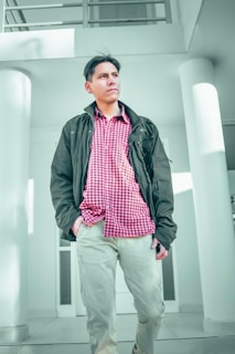 A person stands confidently in a modern architectural setting, wearing a red checkered shirt, a dark jacket, and light pants. The background features white columns and clean lines, suggesting a contemporary building interior.
