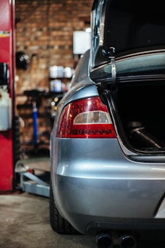 A silver car is parked inside a garage or workshop. The focus is on the rear side, with the trunk open. The background is blurred, displaying a brick wall and some shelves with small objects. The environment has an industrial feel.