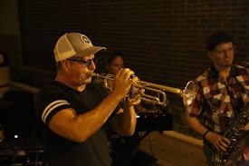 A man wearing sunglasses and a cap is playing a trumpet passionately, with another person playing a saxophone beside him. The setting appears to be outdoors at night with a brick wall in the background.