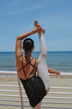 A ballerina in a black leotard and white tights is performing a stretch on a railing by the beach, with one leg raised high above her head. The background features a clear blue sky and calm ocean waves.