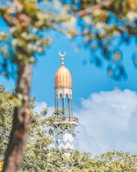 A tall mosque minaret with an ornate dome and crescent atop is framed by lush green foliage and set against a bright blue sky with scattered clouds.