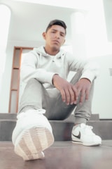 A young person sitting on stairs in a casual pose, dressed in a white hoodie, gray sweatpants, and white sneakers. The focus is on the sneaker in the foreground. The lighting is bright and soft, contributing to the overall casual and relaxed atmosphere of the scene.