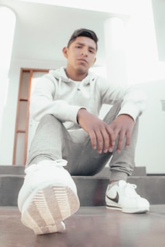 A young person sitting on stairs in a casual pose, dressed in a white hoodie, gray sweatpants, and white sneakers. The focus is on the sneaker in the foreground. The lighting is bright and soft, contributing to the overall casual and relaxed atmosphere of the scene.
