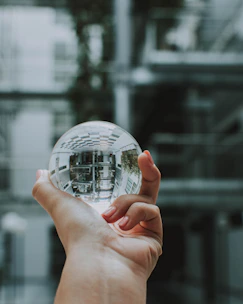 A hand holds a clear, reflective sphere, capturing an upside-down image of the surrounding architecture. The background consists of soft-focus modern building structures with a mix of metal and glass elements.