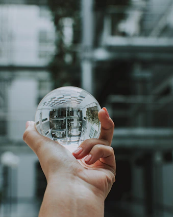 A hand holds a clear, reflective sphere, capturing an upside-down image of the surrounding architecture. The background consists of soft-focus modern building structures with a mix of metal and glass elements.