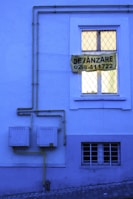 A blue-toned building facade with a window featuring a double-paned style and a yellow sign that reads 'DE VÂNZARE 0268-411722'. Yellow gas pipes run along the wall into two square utility boxes.