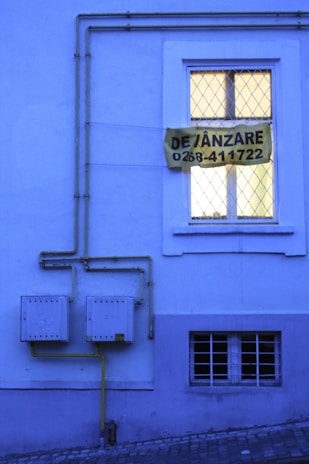 A blue-toned building facade with a window featuring a double-paned style and a yellow sign that reads 'DE VÂNZARE 0268-411722'. Yellow gas pipes run along the wall into two square utility boxes.