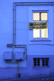 A blue-toned building facade with a window featuring a double-paned style and a yellow sign that reads 'DE VÂNZARE 0268-411722'. Yellow gas pipes run along the wall into two square utility boxes.
