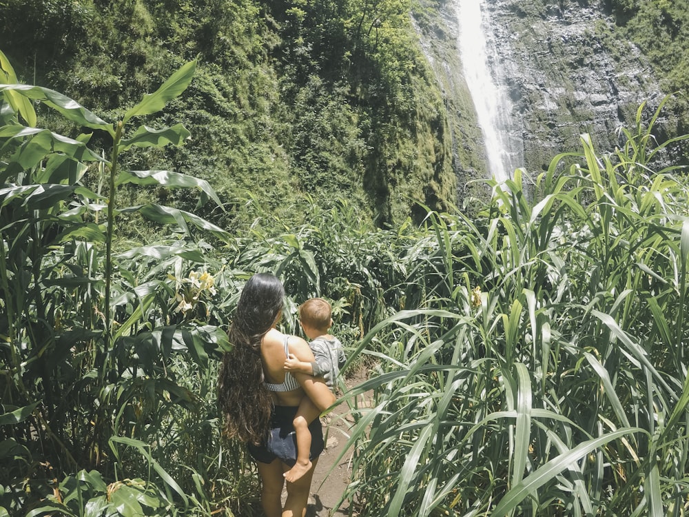 woman holding baby walking on plant field front of waterfal