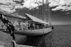 A large sailboat docked at a marina, featuring multiple sails tied down. The boat is equipped with various rigging and maritime equipment. The sky is partly cloudy, and the water is calm. An American flag is visible at the stern of the boat.