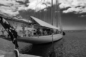 A large sailboat docked at a marina, featuring multiple sails tied down. The boat is equipped with various rigging and maritime equipment. The sky is partly cloudy, and the water is calm. An American flag is visible at the stern of the boat.