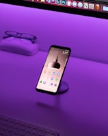 A smartphone is displayed on a stand with its screen on, showing various app icons. It's situated on a desk with a vibrant purple lighting ambiance. In the background, a pair of glasses and a case sit on the left side, and part of a keyboard is visible at the bottom.