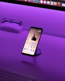 A smartphone is displayed on a stand with its screen on, showing various app icons. It's situated on a desk with a vibrant purple lighting ambiance. In the background, a pair of glasses and a case sit on the left side, and part of a keyboard is visible at the bottom.