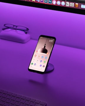A smartphone is displayed on a stand with its screen on, showing various app icons. It's situated on a desk with a vibrant purple lighting ambiance. In the background, a pair of glasses and a case sit on the left side, and part of a keyboard is visible at the bottom.