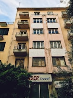 A multi-story building with a worn and weathered exterior. The facade is beige with patches of peeling paint, and several windows and small balconies are visible. There is a sign at the ground level that reads 'travel' accompanied by a partially obscured red word. Bushes are growing next to the building, and neighboring structures with similar designs are visible on one side.