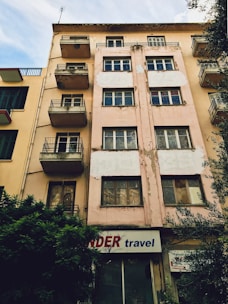 A multi-story building with a worn and weathered exterior. The facade is beige with patches of peeling paint, and several windows and small balconies are visible. There is a sign at the ground level that reads 'travel' accompanied by a partially obscured red word. Bushes are growing next to the building, and neighboring structures with similar designs are visible on one side.