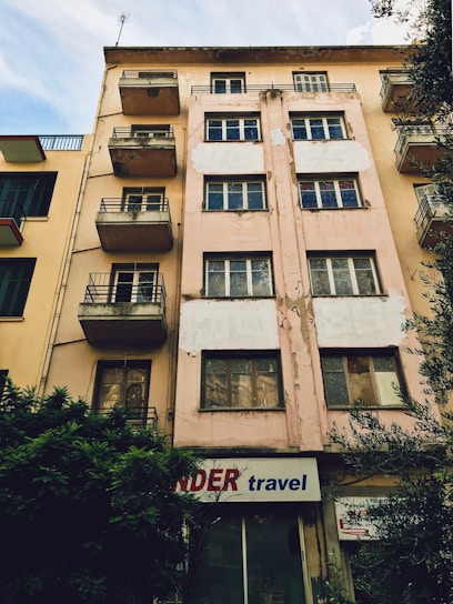 A multi-story building with a worn and weathered exterior. The facade is beige with patches of peeling paint, and several windows and small balconies are visible. There is a sign at the ground level that reads 'travel' accompanied by a partially obscured red word. Bushes are growing next to the building, and neighboring structures with similar designs are visible on one side.