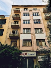 A multi-story building with a worn and weathered exterior. The facade is beige with patches of peeling paint, and several windows and small balconies are visible. There is a sign at the ground level that reads 'travel' accompanied by a partially obscured red word. Bushes are growing next to the building, and neighboring structures with similar designs are visible on one side.