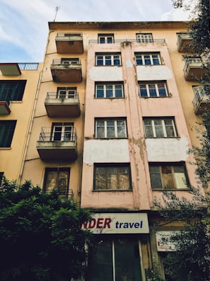 A multi-story building with a worn and weathered exterior. The facade is beige with patches of peeling paint, and several windows and small balconies are visible. There is a sign at the ground level that reads 'travel' accompanied by a partially obscured red word. Bushes are growing next to the building, and neighboring structures with similar designs are visible on one side.