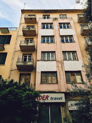 A multi-story building with a worn and weathered exterior. The facade is beige with patches of peeling paint, and several windows and small balconies are visible. There is a sign at the ground level that reads 'travel' accompanied by a partially obscured red word. Bushes are growing next to the building, and neighboring structures with similar designs are visible on one side.