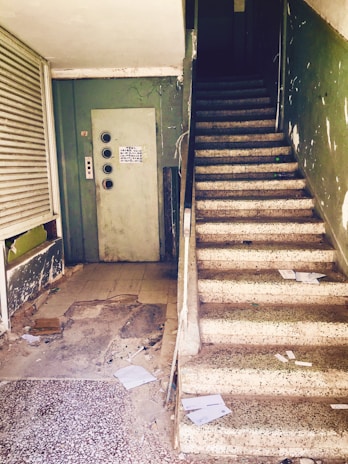 An old, worn-out stairwell in a building, with a dark green wall and beige stairs covered in scattered papers and trash. The area appears neglected and dirty, with cracked surfaces and peeling paint. An elevator door with buttons is visible on the left side, showing signs of decay.