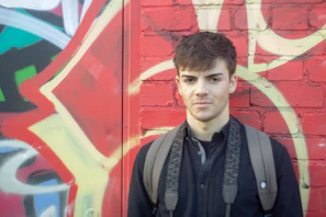 Close-up of a young man wearing a black Rudrawears graphic tee with a fierce red print, standing against a gritty urban wall.