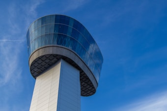 A modern air traffic control tower with a cylindrical design composed of glass and steel panels. The structure is set against a clear blue sky, emphasizing its elegant and functional architecture.