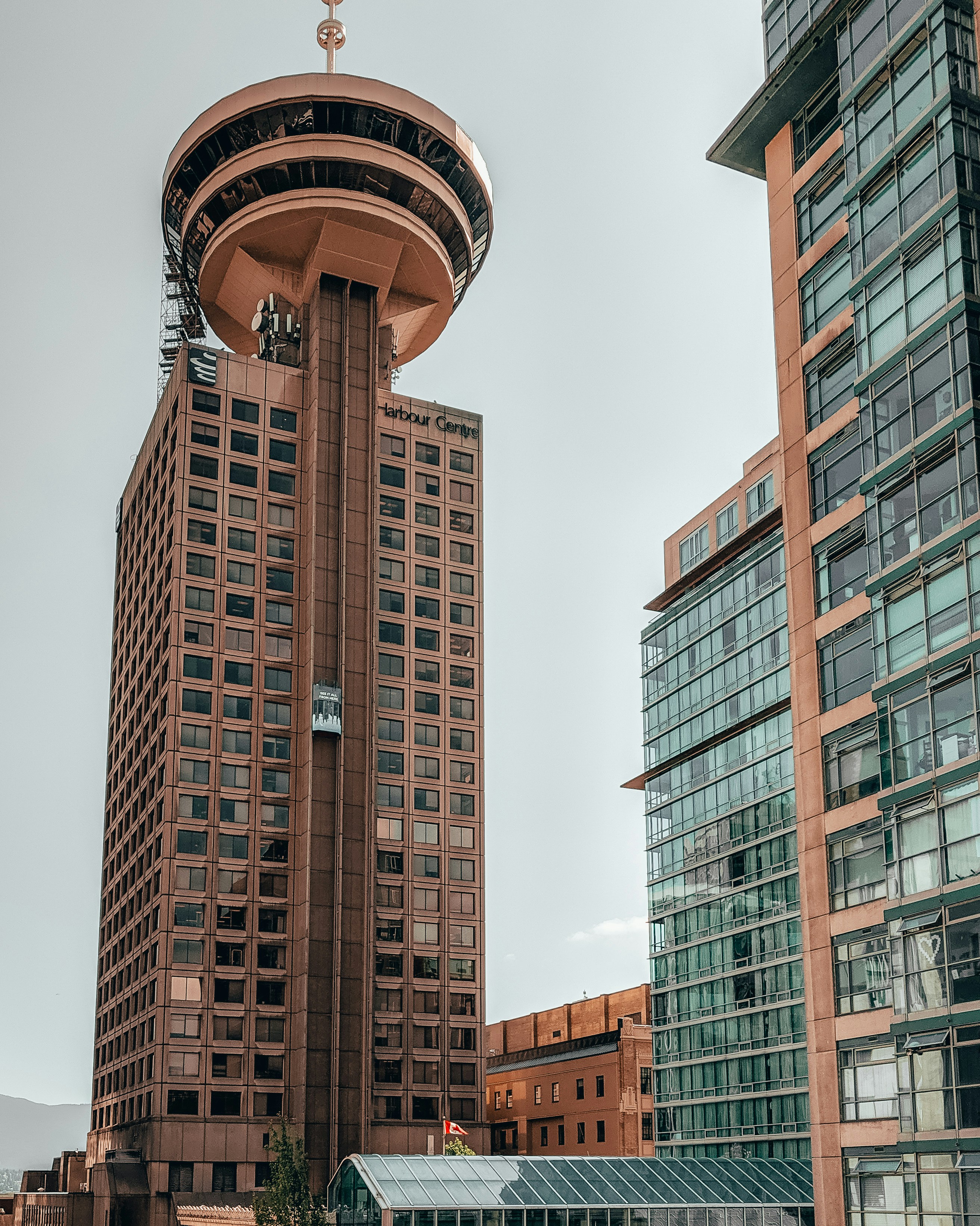A photo of The Vancouver Lookout in Vancouver, Canada. The cylindrical tower with observation windows sits atop a building in the downtown area, with mountains and the harbor visible in the background. (Photo by Marco Tjokro on Unsplash)